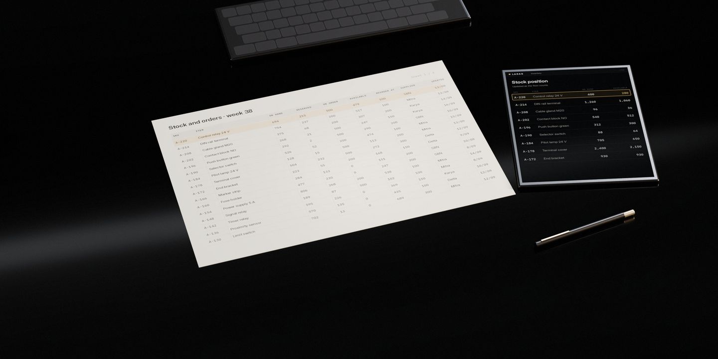 A wide printed stock sheet on a dark stone desk, a keyboard behind it, and a tablet showing the same rows live, one of them marked