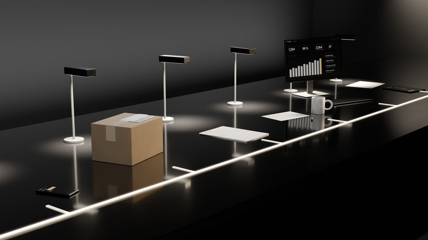 One long dark stone desk with six workplaces, each under its own lamp — a phone, a parcel, a stack of invoices, a person’s place with a mug, a screen and keyboard, a tablet — first with slips of paper scattered along the desk between them and the screens dark, then with one lit line running through every workplace and every screen on